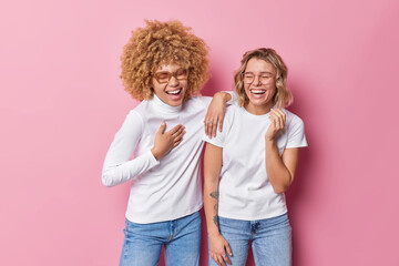 Overjoyed women laugh happily have fun keep eyes closed smile broadly dressed in casual clothes isolated over pink background. Two female friends foolish around hear funny story stand indoor