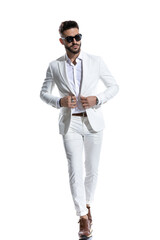 elegant unshaved guy with sunglasses buttoning white suit and walking