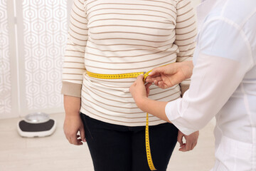 Nutritionist measuring overweight woman's waist with tape in clinic, closeup