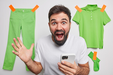 Surprised shocked bearded young man holds mobile phone reacts on amazing news exclaims loudly dressed in casual basic t shirt poses indoor with green clothes hanging behind sells clothing online