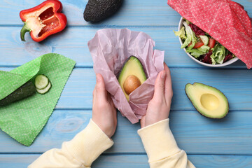 Woman packing half of fresh avocado into beeswax food wrap at light blue wooden table, top view - obrazy, fototapety, plakaty