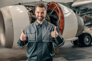 Cheerful man aircraft maintenance engineer giving thumbs up and smiling while standing near airplane