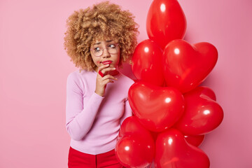 Frustrated curly haired crying woman feels unloved looks sadly away holds bunch of inflated red heart shaped balloons thinks about finding true love on Valentines Day dressed in casual clothes