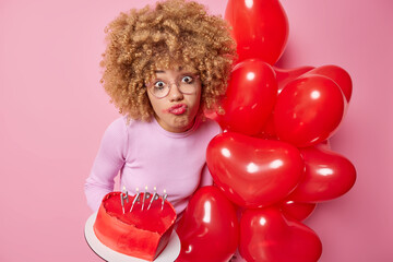 Upset curly haired young woman feels lonely on Valentines Day holds bunch of heart shaped balloons and delicious cake wears transparent glasses has spoiled makeup poses against pink background