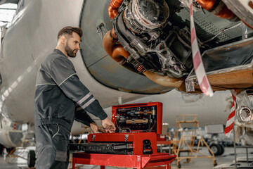 Male worker maintenance technician putting instrument in tool box while standing near airplane at...