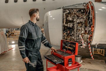 Bearded man airline maintenance technician holding wrench and looking at airplane while standing...