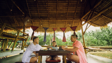 The happy family eat and drink tasty beverages spending time in local floating cafe on water. Father, mother and daughter having breakfast outdoors. Man, woman and a child in a cafe. Tropical view
