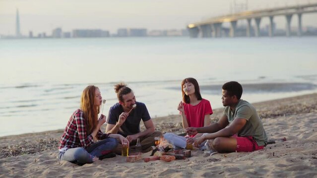 Four multi-ethnic young friends sitting around campfire on beach, roasting and eating marshmallows on sticks and talking on summer weekend. Diverse people spending vacation together