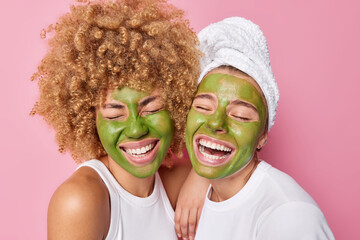 Horizontal shot of happy women applies green facial mask made of natural ingredients stand closely...