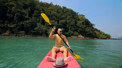 Young man with sunglasses and hat rows pink plastic canoe along sea against green hilly islands with wild jungles. Traveling to tropical countries. Strong guy is sailing on kayak in ocean, front view.