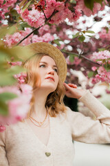 Fototapeta premium Young stylish woman standing in sakura park and enjoy beauty of pink. Spring woman in cherry flower bloom concept. Beauty blonde woman with long hair outdoors. Cherry blossoms. Close up portrait.