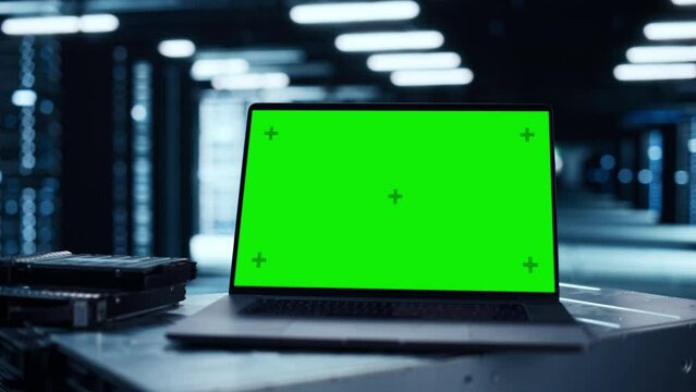 Laptop Computer with Green Screen Chroma Key Mock Up Display Stands on a Table. In the Background Modern Data Center With Rows of Server Racks. Concept for Cloud Computing and Cyber Security.