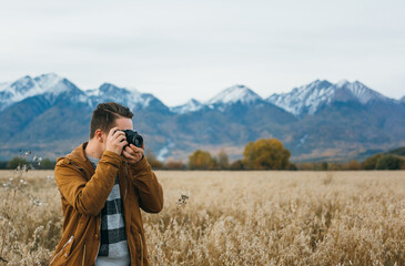 The guy is a photographer in the field, against the background of mountains, takes a picture on an old retro film camera. Man walks in the fall in casual clothes. Concept lifestyle millennial hipster - Powered by Adobe
