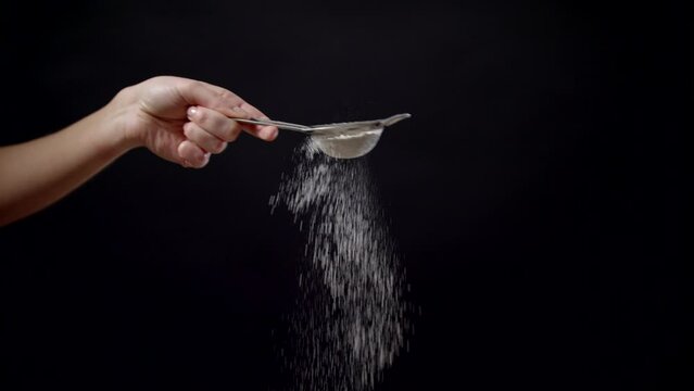 Closeup Slowly Flour Sifting With Sieve, Female Chef Sifts Powdered Sugar Or Flour In Sieve On Black Background. Homemade Dough, Bread, Pizza, Cupcake Preparation. Young Hands Sift Flour