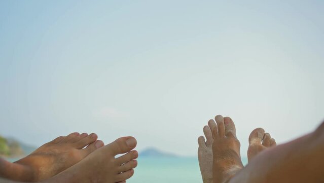 Bare feet of young man and woman couple wiggle lying on beach near calm azure ocean at exotic resort close first point view. Pov of leg of a couple of men and women lying on a tropical sandy beach.