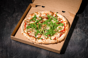 Pizza in paper box on black stone background. Italian traditional food. Delicious pizza with pear, parmigiano-reggiano cheese and rucola. Restaurant menu, food delivery and take-out. Copy space