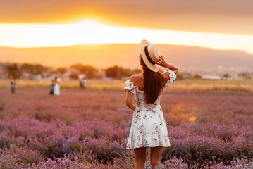 A young beautiful girl in a delicate dress and hat walks through a beautiful field of lavender and enjoys the fragrance of flowers. Rest and beautiful nature. Lavender blooming and flower picking.