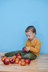 Fototapeta premium a boy of 2 years sits on a wooden table and plays with apples and tangerines, he closes his eyes and plays a game.