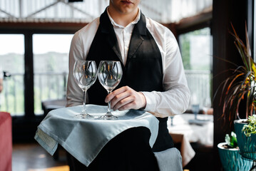 A young waiter stands with wine glasses in a stylish modern restaurant. Table service in the restaurant. Haute cuisine, service and maintenance. Close-up.