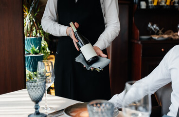 A young sommelier in a stylish uniform demonstrates and offers the client fine wine in the restaurant. Customer service. Table setting in a fine restaurant.