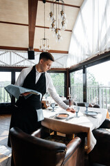 A young male waiter in a stylish uniform is engaged in serving the table in a beautiful gourmet...