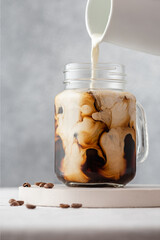 Pouring milk or cream into the iced coffee in a glass jar on gray background. Cold refreshment summer drink