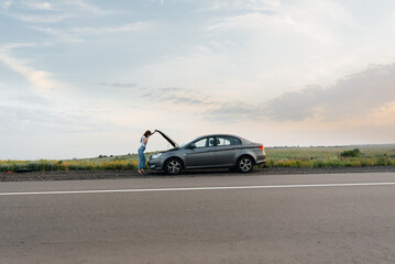 A young girl stands near a broken-down car in the middle of the highway during sunset and tries to call for help on the phone. Waiting for help. Car service. Car breakdown on road.