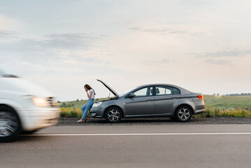 Obraz premium A young girl stands near a broken car in the middle of the highway during sunset and tries to call for help on the phone and start the car. Waiting for help. Car service. Car breakdown on the road.