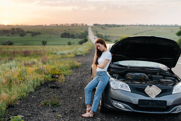 A frustrated young girl stands near a broken-down car in the middle of the highway during sunset....