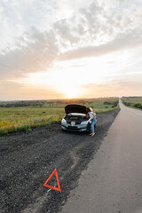 A young girl stands near a broken car in the middle of the highway during sunset and tries to repair it. Troubleshooting the problem. Waiting for help. Car service. Car breakdown on road..