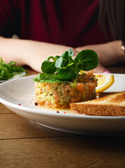Salmon tartare with avocado, arugula, spinach, bread chips and lemon. On a plate, on a wooden background. A woman in a red dress eats salmon tartare. Hands, fork close-up. Healthy healthy food. Raw