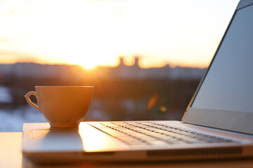Coffee cup on laptop against the window with sunshine. Cozy workplace in home office, concept of remote work and earning online