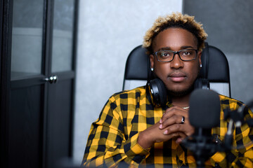 Influential African-American man with glasses confidently looks into the camera next to the microphone in the studio. A black guy keeps his hands folded, sitting comfortably in a chair.