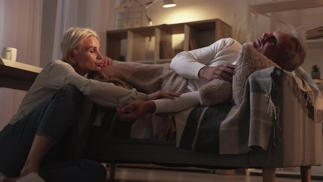 Home disease. Sick man. Family health care. Worried middle-aged woman patting hand her husband laying on sofa with fever in light shadow room interior.