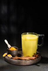 A glass cup of golden milk with turmeric powder and fresh turmeric root and anise and cinnamon on black. The concept of spices and herbs. Ayurvedic drink on a light background. Space for text.