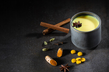 A glass cup of golden milk with turmeric powder and fresh turmeric root and anise and cinnamon on black. The concept of spices and herbs. Ayurvedic drink on a light background. Space for text.