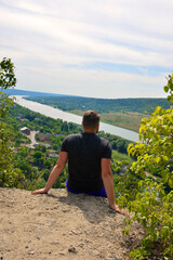 a man sits on the edge of a cliff, rests after a long climb and admires the beautiful landscape: river, forest and mountains