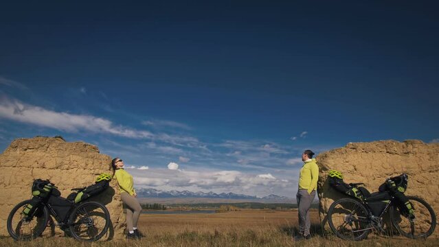 The man and woman travel on mixed terrain cycle bike touring with bikepacking. The two people journey with bicycle bags. Sport sportswear in green black colors. Mountain snow capped, stone arch.