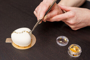A woman decorates white mousse cake on gold substrate with confectionery gold with tweezers....