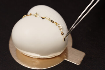 White mousse cake on gold substrate is decorated with gold with tongs. Selective focus.