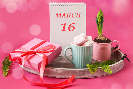 Calendar for March 16: a notepad with the inscription 16 March in English, blooming hyacinth, air marshmallow in a cup, a gift tied with a pink ribbon on a pink background, bokeh