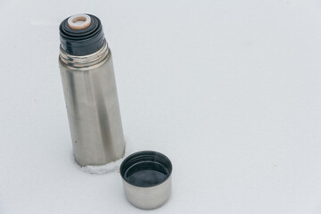 Thermos with a hot drink and a cup on a background of snow. Tourist utensils. Drink hot tea in winter to keep warm.