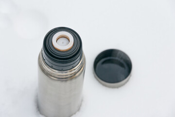 Thermos with a hot drink and a cup on a background of snow. Tourist utensils. Drink hot tea in winter to keep warm.