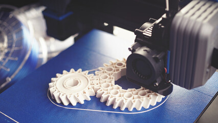 white helical gears produced by small black FDM-printer on blue build plate. clean bright technical sceney in wide format with filament rolls in background. excessive selective focus.  - obrazy, fototapety, plakaty
