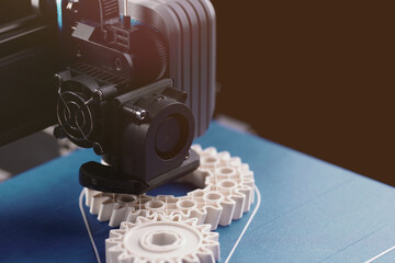 FDM-3D-printer with blue print platform manufactures white helical gears from plastic filament in...