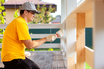 A man in a bright yellow T-shirt and cap holds a brush in his hand and paints the boards on the porch of the house with white paint