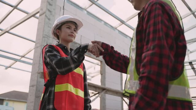 Happy Asian Female Architect And Male Contractor Wear White Safety Hard Hats Standing Shaking Hands For Business Partnership In The Construction Site. Civil Engineer And Specialist Builder Handshake. 