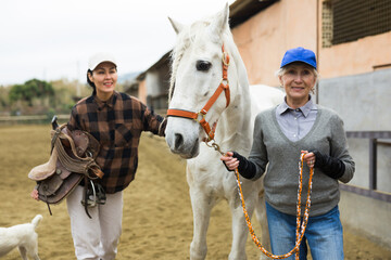 Women ranchers preparing white horse for ride. Senior European woman leading horse, Asian woman...