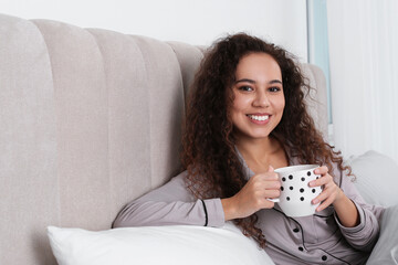 Beautiful African American woman with cup of drink in bed at home