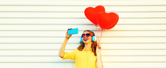 Portrait of happy smiling cheerful young woman taking selfie by smartphone with red heart shaped balloons listening to music on white background